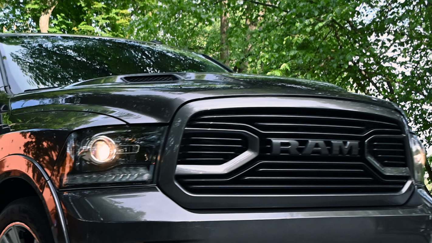 A close-up view of a black RAM truck's front grille and headlights, set against green foliage.