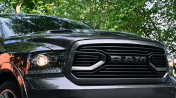 A close-up view of a black RAM truck's front grille and headlights, set against green foliage.