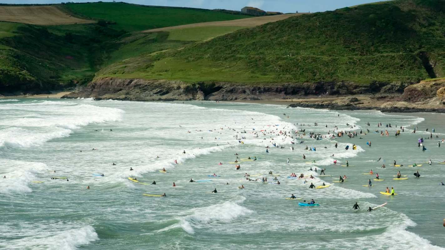 A crowded beach scene with numerous surfers riding waves and green hills in the background.