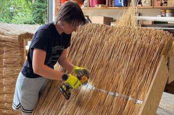 A person using a power drill to fasten metal strips onto a large bundle of straw in a workshop setting.