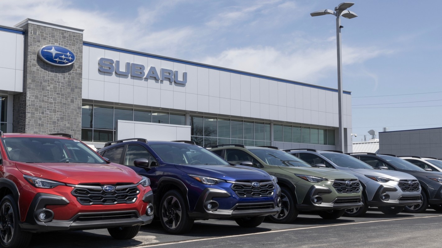 A row of Subaru SUVs parked in front of a Subaru dealership under a clear blue sky.