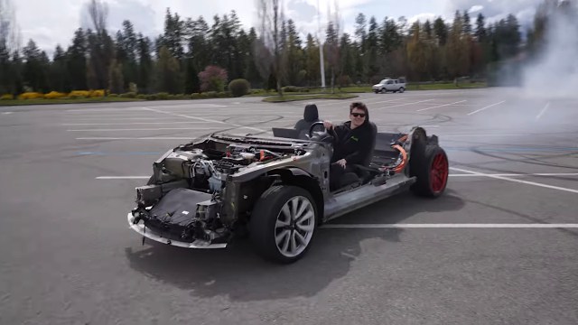 A person is driving a stripped-down, modified car in a parking lot, surrounded by trees.