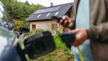 An electric vehicle is plugged in outside a home with solar panels set against a wooded backdrop.