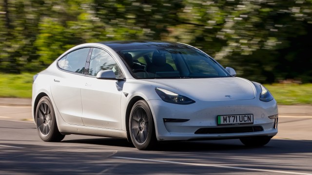 A white Tesla Model 3 driving on a scenic road with blurred green foliage in the background.