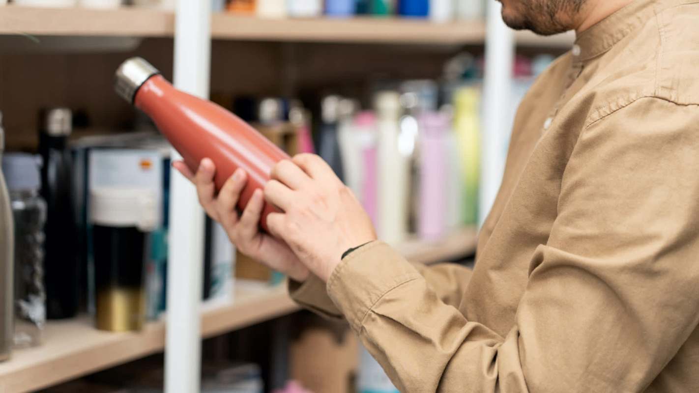A person examines a red metal water bottle while standing in front of a shelf filled with various containers.