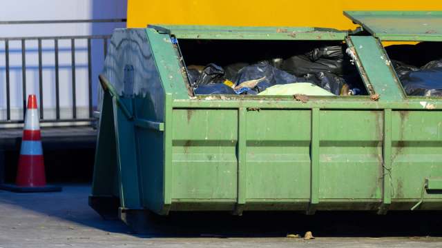 A green dumpster filled with black trash bags sits in a parking area beside a traffic cone.