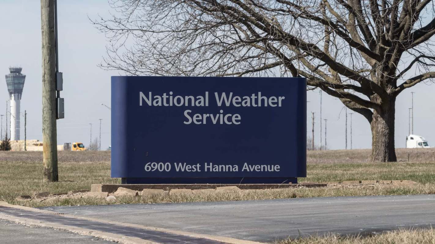 A sign for the National Weather Service, located at 6900 West Hanna Avenue, with an airport control tower visible in the background.