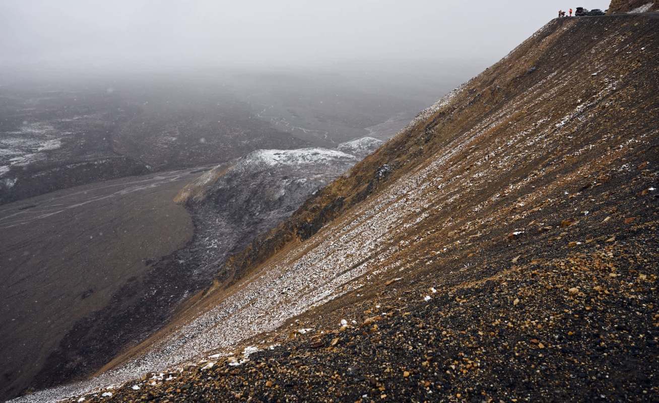 A steep, rocky slope created by a landslide, covered in snow and overlooking a gray, misty landscape with distant hills.