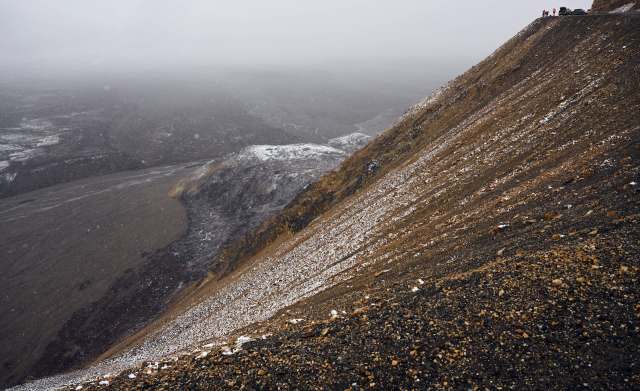 A steep, rocky slope created by a landslide, covered in snow and overlooking a gray, misty landscape with distant hills.