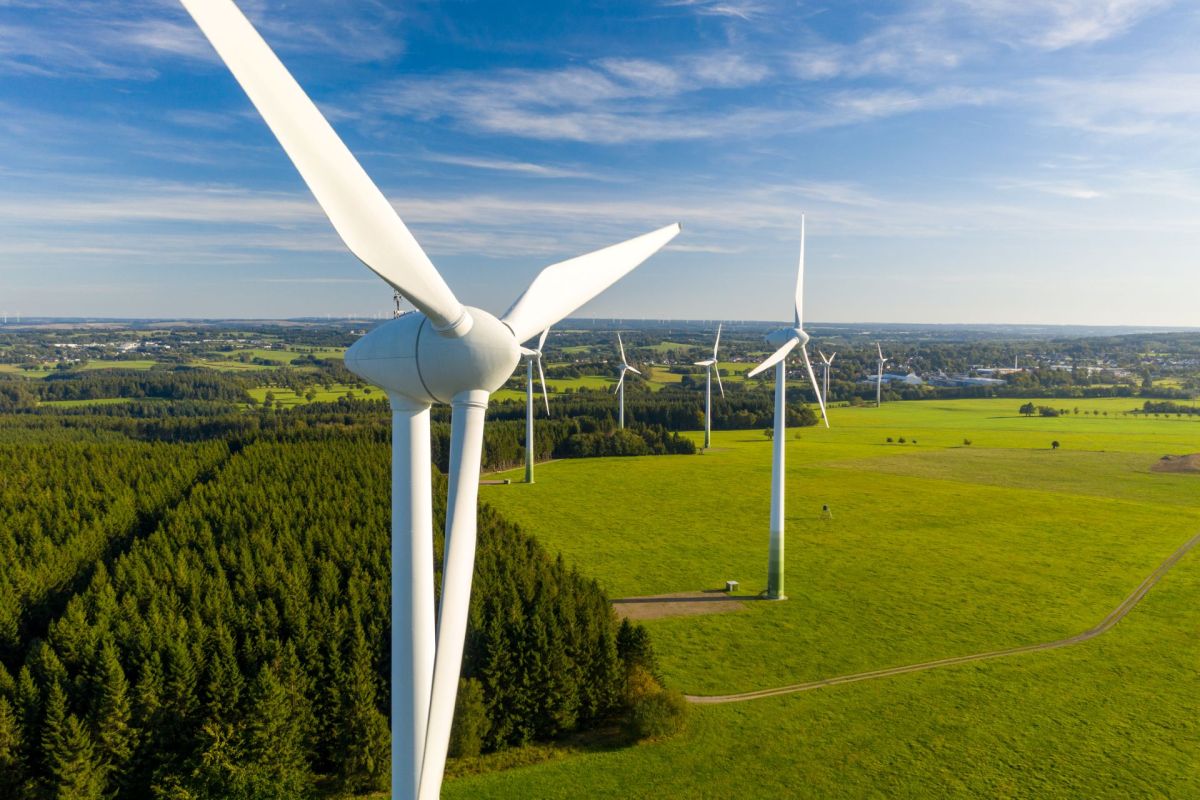 Aerial view of wind turbines in a green landscape with fields and forests under a clear blue sky.