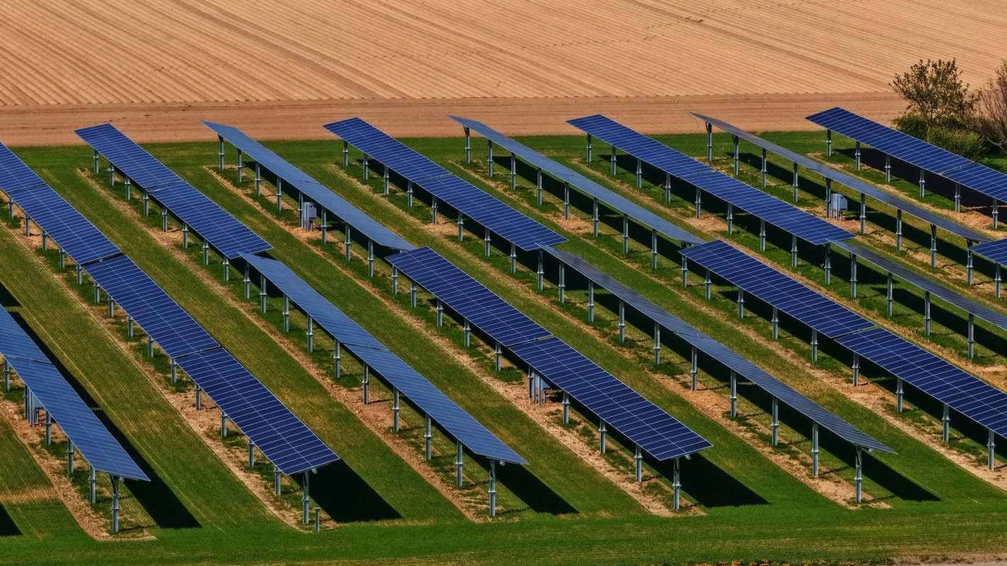 Aerial view of a solar farm featuring rows of solar panels on green grass, with farmland in the background.