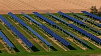 Aerial view of a solar farm featuring rows of solar panels on green grass, with farmland in the background.