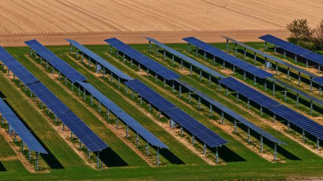 Aerial view of a solar farm featuring rows of solar panels on green grass, with farmland in the background.
