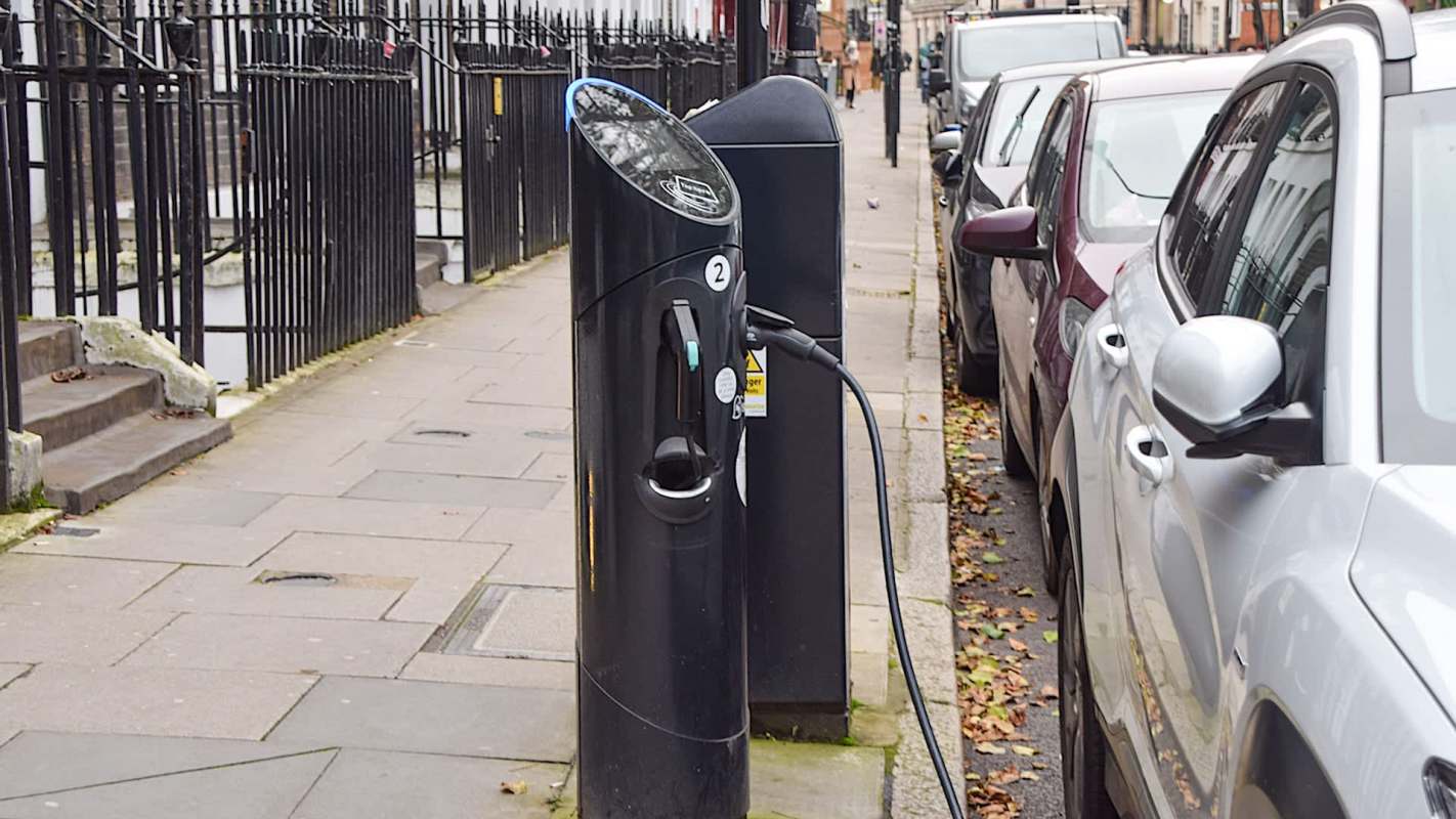An electric vehicle charging station located on a sidewalk, surrounded by parked cars.