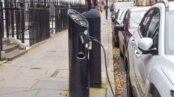 An electric vehicle charging station located on a sidewalk, surrounded by parked cars.