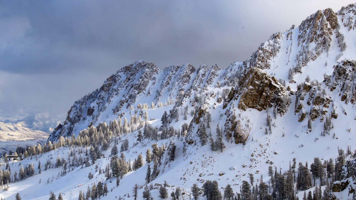 Snow-covered mountains rise against a cloudy sky, with evergreen trees scattered along the slopes.