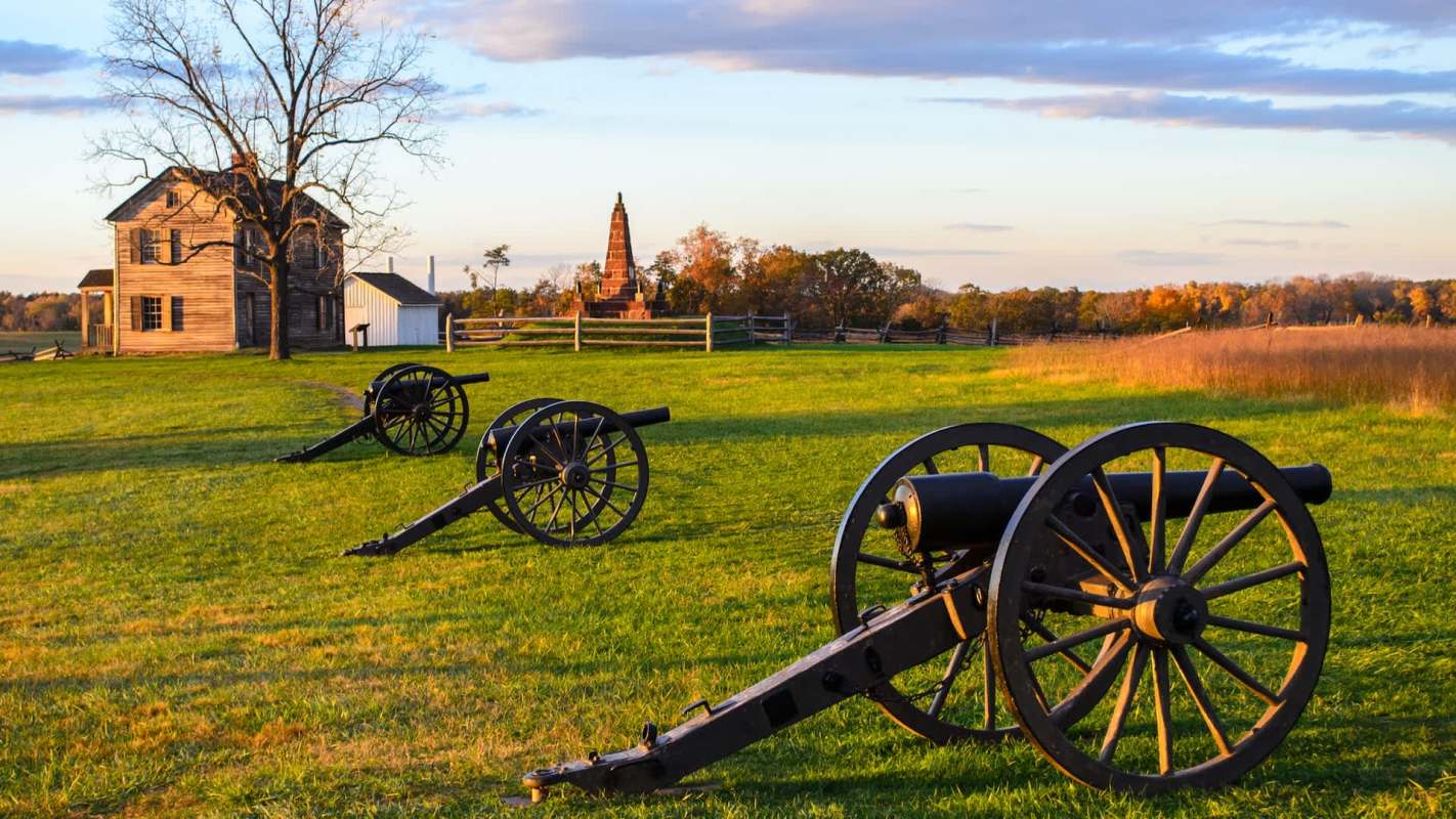 Three historic cannons on green grass with a wooden house and a tall monument in the background under a sunset sky.