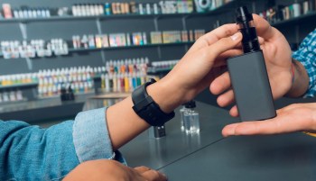 A person hands a vape device to another person in a vape shop with colorful product displays in the background.