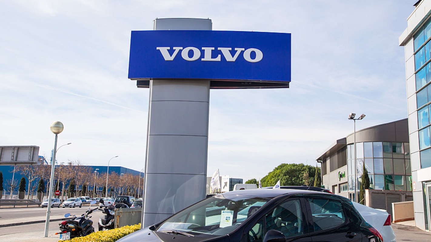 A Volvo dealership sign stands prominently next to parked cars and modern buildings under a clear sky.