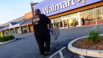 A person pushing a shopping cart enters Walmart at sunset, with other shoppers nearby.