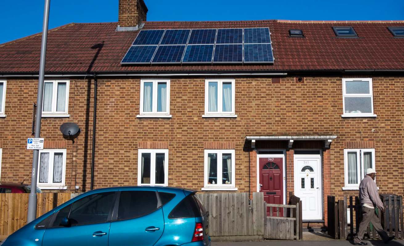A brick townhouse with solar panels on the roof, featuring two doors and a passerby walking on the sidewalk.