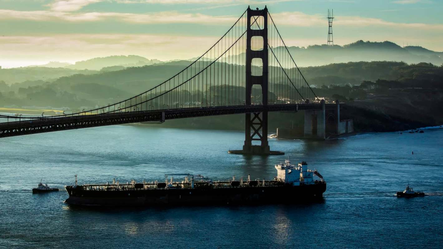 A cargo ship passes under the Golden Gate Bridge, surrounded by misty hills and calm waters.