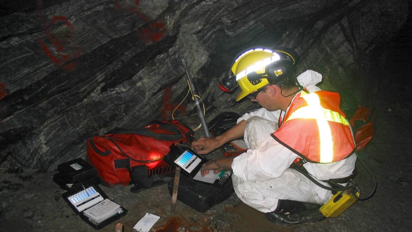 A miner in protective gear conducts field research in a dark underground environment.