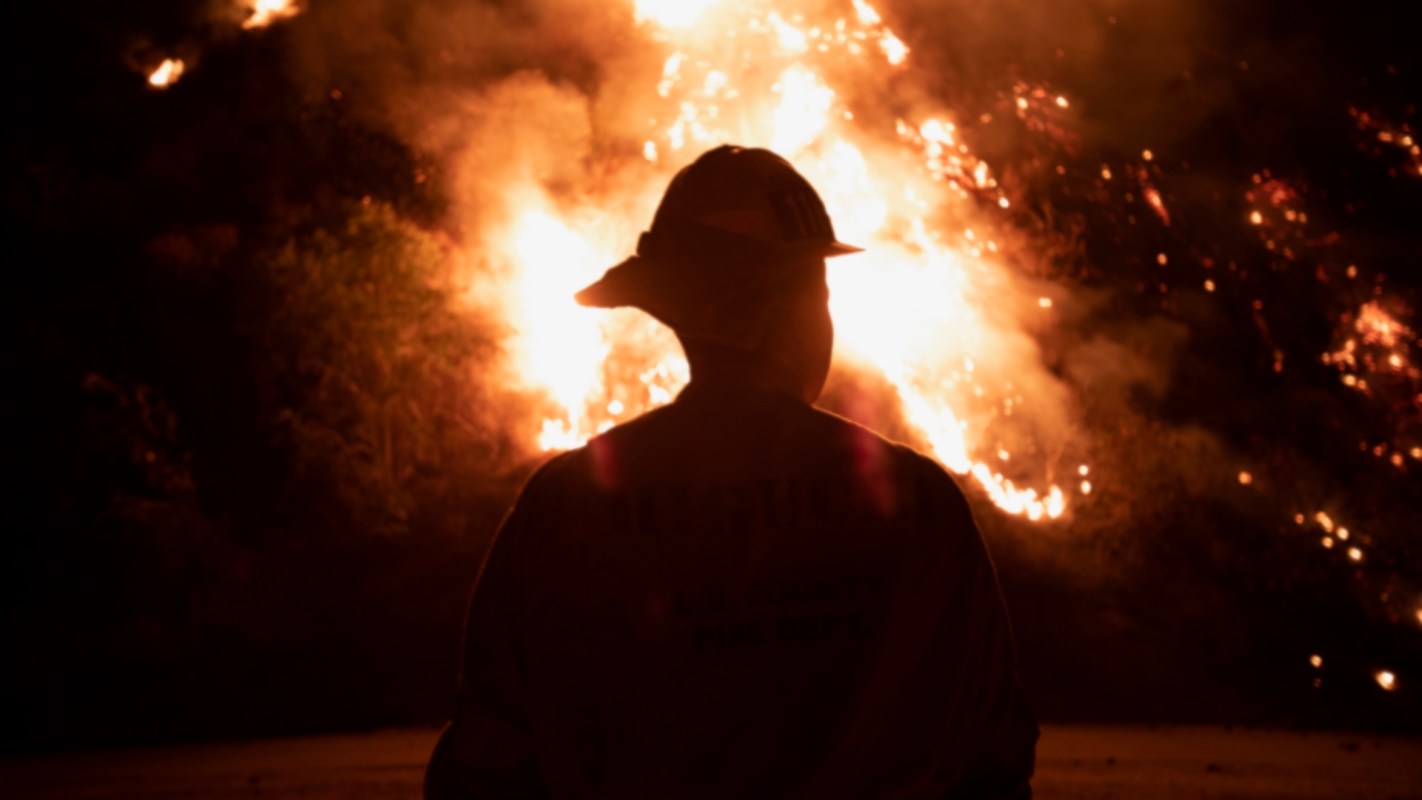 A firefighter standing in front of a wildfire at night.