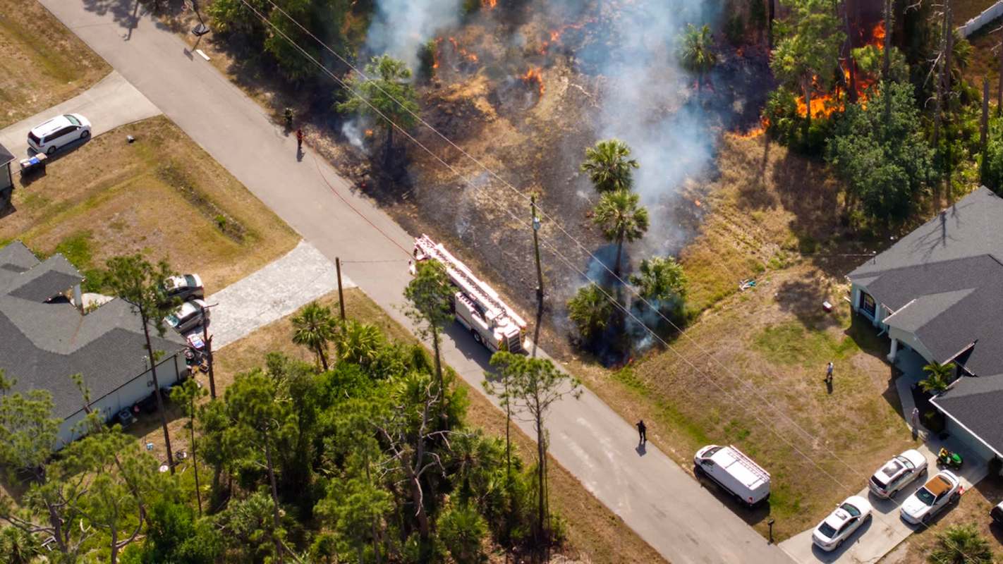 Aerial view of a fire truck responding to a brush fire near residential homes with smoke and flames visible.