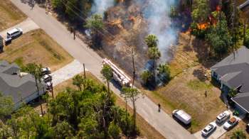 Aerial view of a fire truck responding to a brush fire near residential homes with smoke and flames visible.