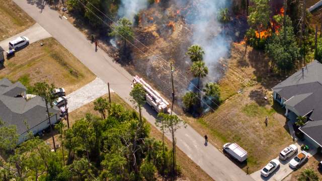Aerial view of a fire truck responding to a brush fire near residential homes with smoke and flames visible.