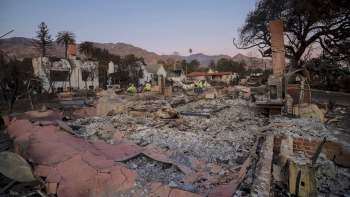 A devastated neighborhood with burnt homes and debris scattered, set against mountains and a twilight sky.