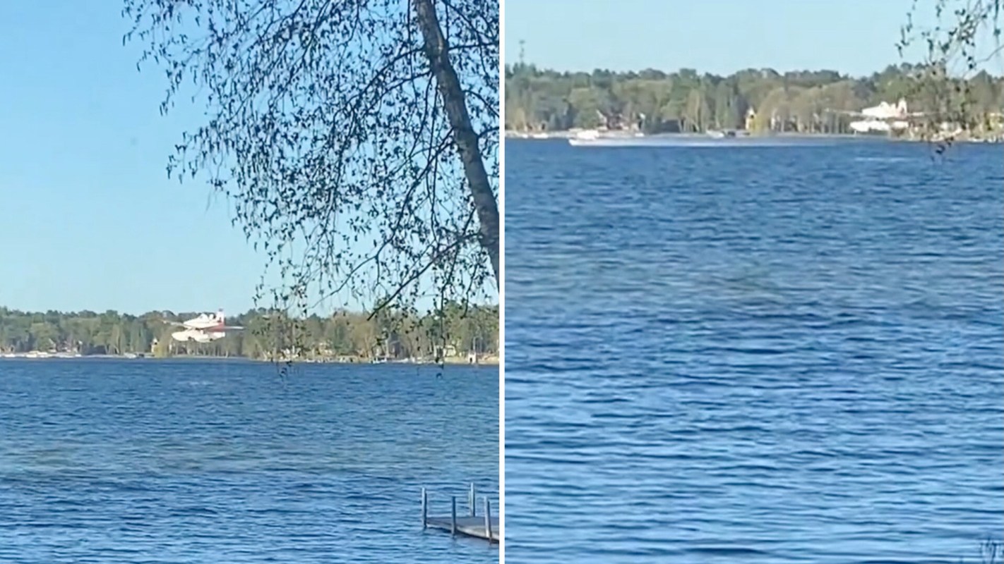 A small airplane flies low above a lake surrounded by trees.