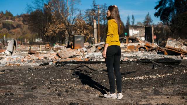 A woman in a yellow sweater stands amid the ruins of a burned area, observing the destruction around her.