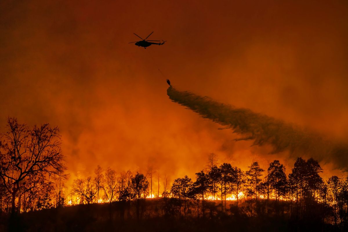 A helicopter battles a wildfire, spraying water over flames and smoke against a dark orange sky.
