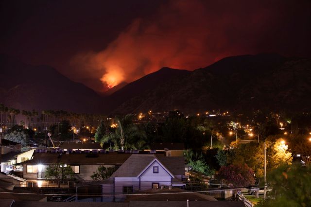 A nighttime view of a valley illuminated by city lights, with smoke and flames rising from the mountains in the background.