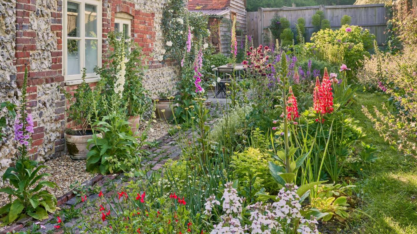 A lush English country garden filled with colorful flowers and plants alongside a brick wall and a small seating area.
