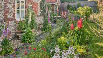 A lush English country garden filled with colorful flowers and plants alongside a brick wall and a small seating area.