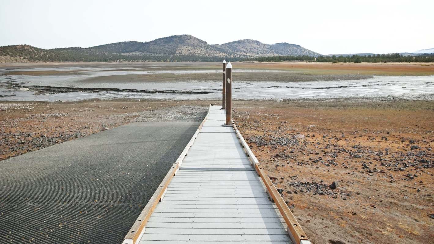 A wooden walkway leads across dry land toward a dry reservoir with distant mountains under a clear sky.