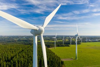 A close-up view of wind turbines over green fields and a forest under a blue sky.