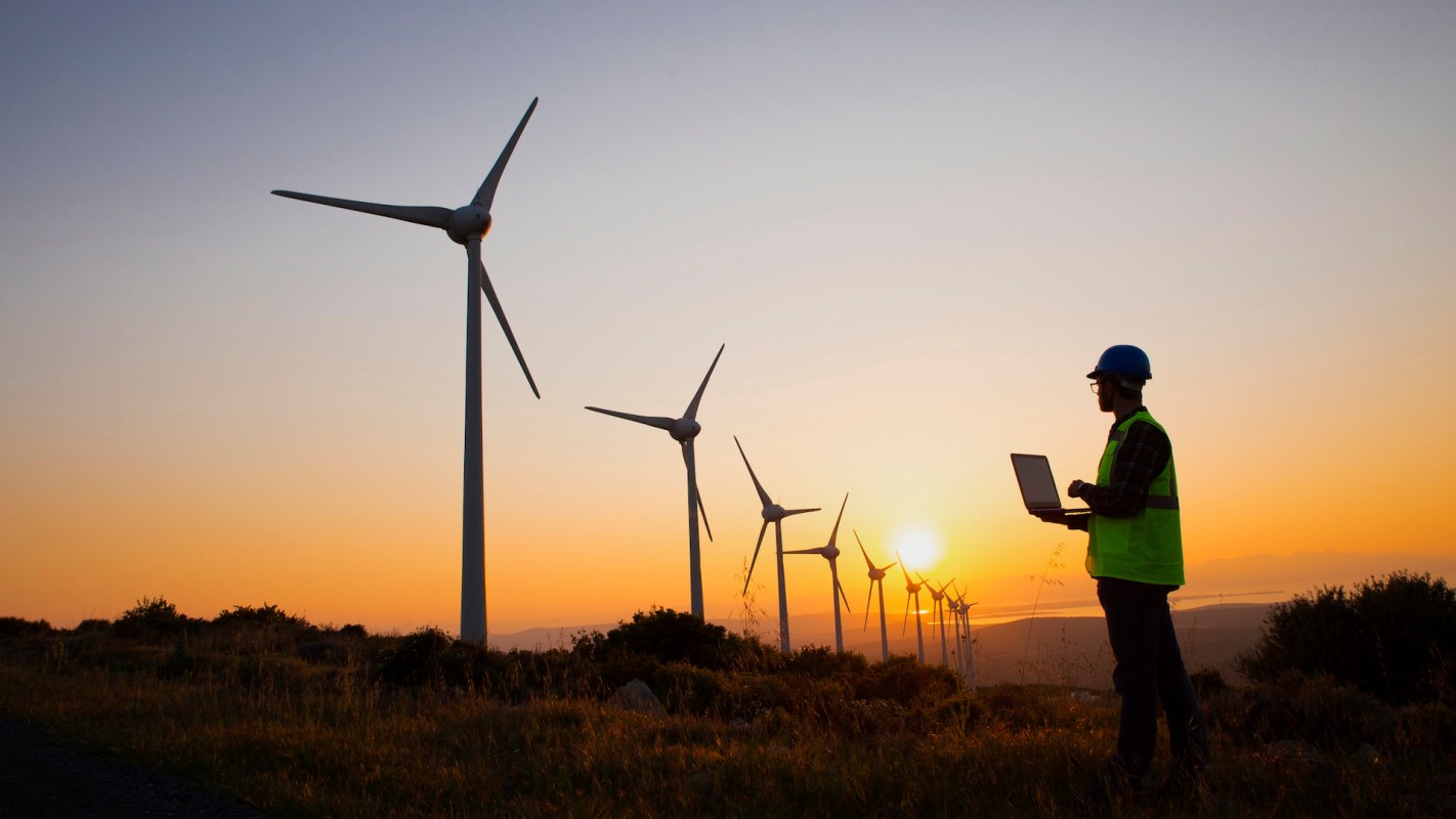 A silhouette of a worker with a laptop standing among wind turbines at sunset.
