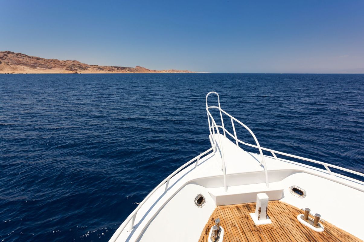 A yacht's bow rises above calm, deep blue waters with a distant shoreline under a clear sky.