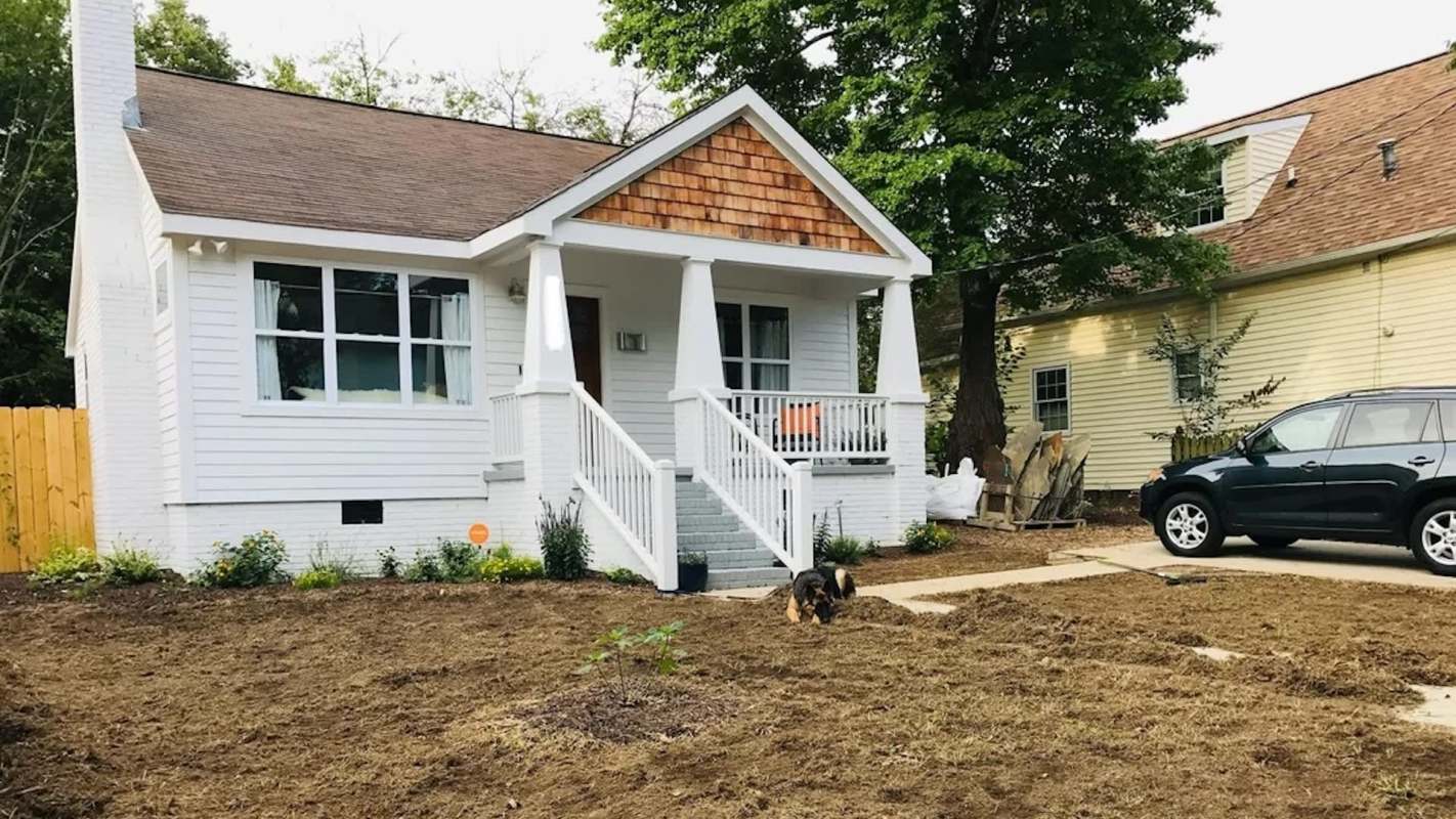 A renovated white cottage with a porch and a dog playing on freshly prepared landscaping.
