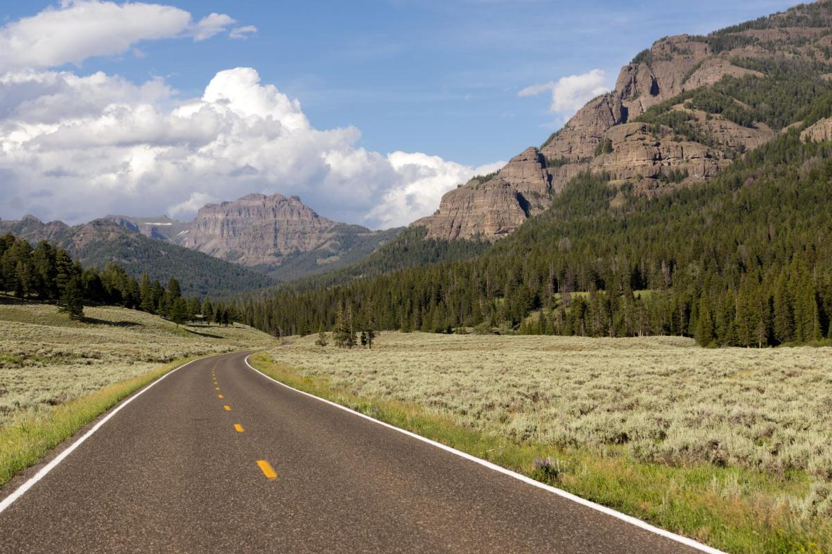 A winding road leads through a green valley with mountains and clouds in the background.