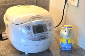 A white rice cooker and a colorful elephant figurine with a yellow shirt and blue hat on a countertop.