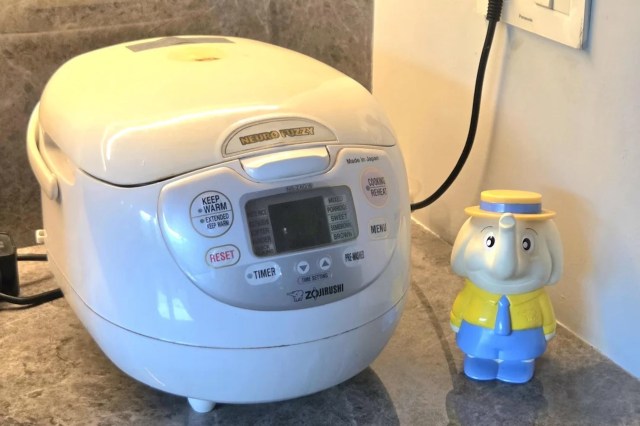 A white rice cooker and a colorful elephant figurine with a yellow shirt and blue hat on a countertop.