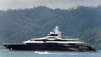 A large luxury yacht anchored near a lush, green hillside with a small speedboat nearby.