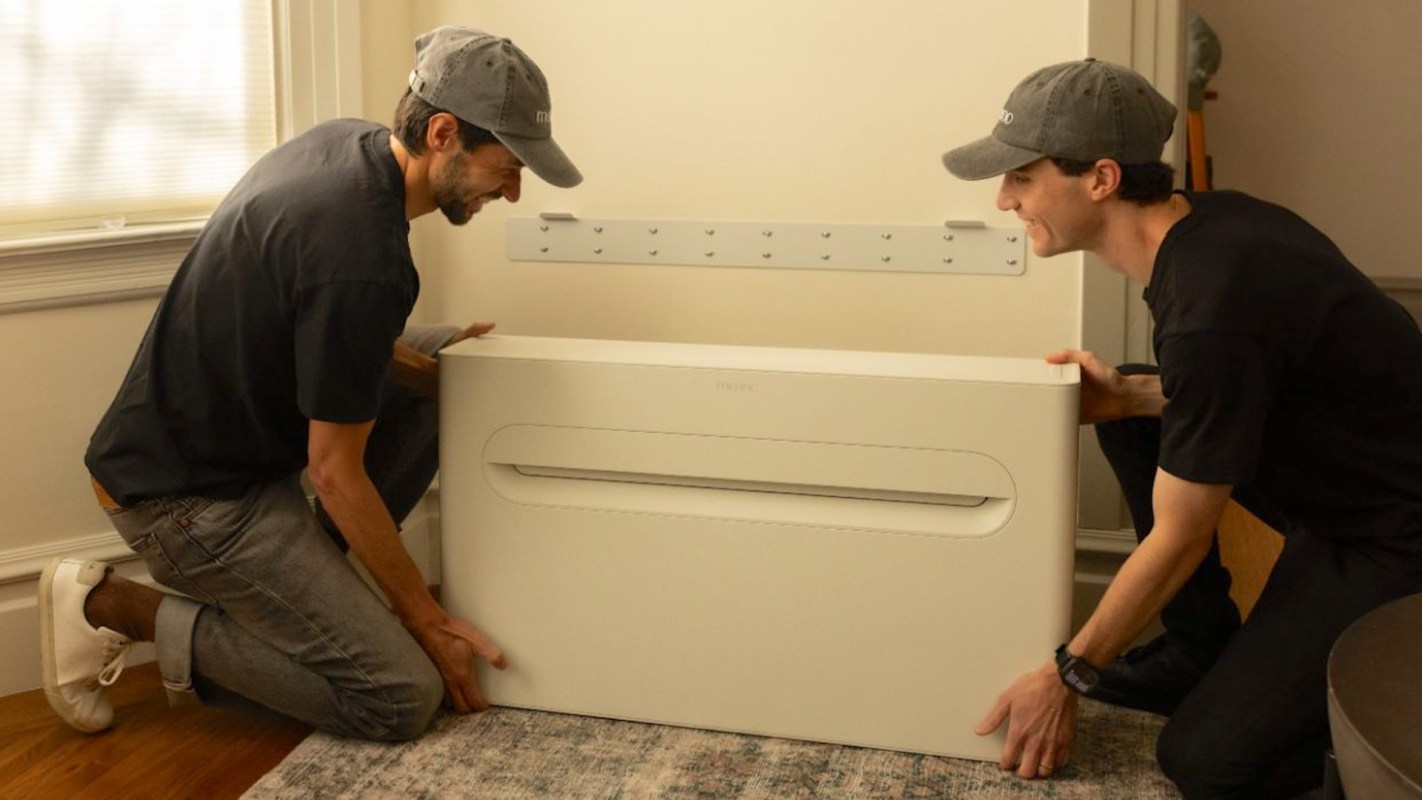 Two men in caps install a Merino Energy heat pump unit against a wall in a living room.