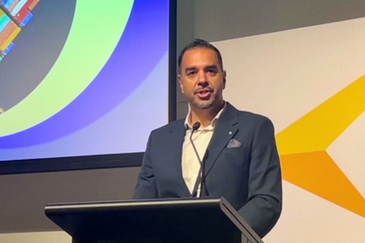 A man in a suit speaks at a podium with a colorful presentation backdrop.