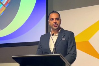 A man in a suit speaks at a podium with a colorful presentation backdrop.